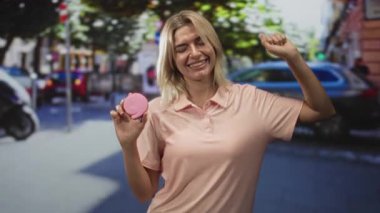Meşgul bir şehir caddesinde kollarını kaldırmış pembe bir makarna tutan kadın, geniş bir gülümseme; neşeli mutluluk.