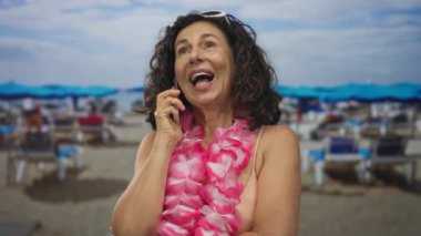 Güneşli bir sahilde telefonla konuşan bir kadın, bikini ve pembe bir lei giyiyor, güneş pansiyonları ve şemsiyelerle çevrili, deniz kenarındaki tropikal tatil havasının tadını çıkarıyor..