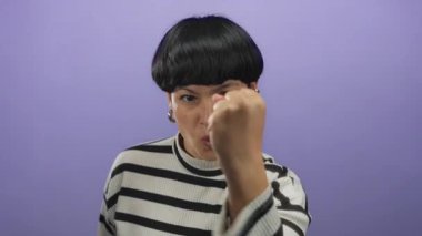 Yumruk kaldırılmış kadın, parlak çizgili süveter giyen, güç, cesaret ve motivasyon uyandıran münferit mor arka plana karşı kararlı görünüyor..