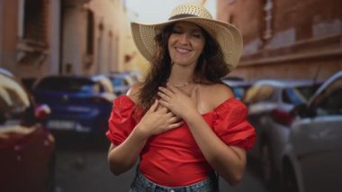 Sokakta göğüs göğüse gülümseyen kadın geniş hasır şapkalı ve kırmızı omuzlu; seyahat neşesi.