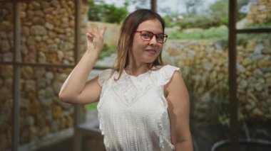 Kadın elleriyle çifte barış işareti yaparken gülümsüyor ve stüdyo duvarlı kırmızı gözlük takıyor; sevinç kutlaması.