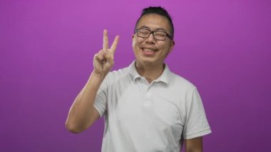 Beyaz polo giyen ve gözlüklü genç bir adam mor bir stüdyoda barış işareti gösterirken elleriyle kalp oluşturuyor..