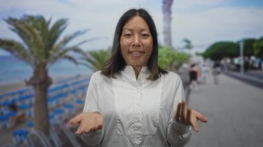 Palmiye ağaçları ve şemsiyelerin yanında güneşli bir plajda omuz silken beyaz gömlekli kadın; dinginlik berraklığı.