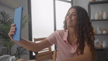 Elinde mavi tabletle stüdyoda selfie çeken bir kadın masadaki çömlek rafları ve bitkilerle görünür. Gençlik sevinci..