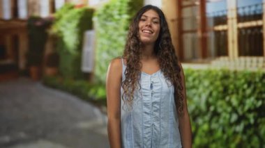 Bir kaldırım taşı caddesinde, yapraklı bir binanın yanında konuşurken el kol hareketi yapan bir kadın; dostça bir açıklama.