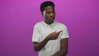 Genç Afrikalı Amerikalı adam mor stüdyoda beyaz polo tişört giydiğini işaret ediyor..