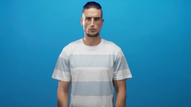 Görünür yüz ve kısa sakallı bir adam kameraya karşı nötr ifade, kollar rahat, çizgili tişört mavi stüdyo; düşünceli yansıma.