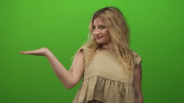 Sarışın, sol elini uzatmış genç bir kadın kameraya gülümserken stüdyo yeşili içinde açık avuç içi sunuyor..