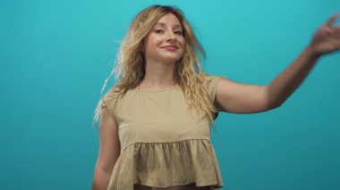 Sarışın kadın gülümsüyor, elleriyle gözlerini kapatıyor ve stüdyoda turkuaz arka plan ve günlük bluzla çıplak kollarını gösteriyor; çekingenlik..