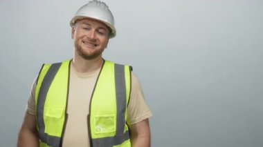 Sert şapkalı ve yüksek görüşlü yelekli bir adam iki elini öne uzatıyor ve soluk mavi stüdyoda gülümsüyor; güvenlik ekibi karşılaması.