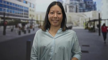 Gülümseyen kadın, açık elini, açık mavi bluzlu bir şehir caddesinde kameraya doğru uzatır..