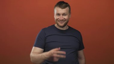 Turuncu stüdyoda el hareketi ve günlük tişört gösteren bir adam kamerayı işaret ediyor..