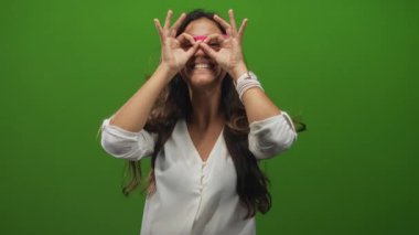 Yeşil bir stüdyoda pembe gözlük takan bir kadın, çıplak ellerle geniş bir gülümseme ile gözlerinin üzerinde dairesel el gözlükleri oluşturur..