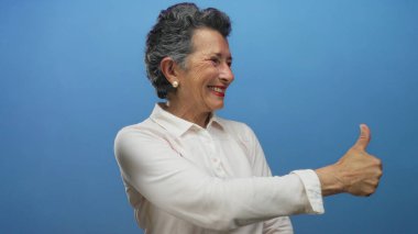 Gri saçlı yaşlı bir kadın mavi arka plana baş parmak sallıyor. Pozitif ve kendinden emin..