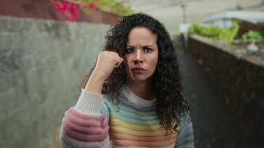 Açık havada, kıvırcık saçlı ve gökkuşağı süveterli bir kadın, yumruğuyla taştan duvar zeminine karşı öfkesini ve kararlılığını canlı kent ortamında çiçeklerle gösteriyor..