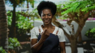 Önlüklü kadın, yemyeşil bir parkta parmaklarıyla bir jest yapıyor. Etrafı yeşillik ve sıcaklıkla çevrili ve gülümsüyor..