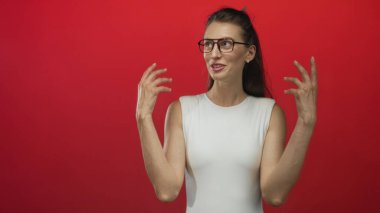 Gözlüklü bir kadın kırmızı stüdyo setinde avuçlarını açıp el hareketlerini gösteriyor, gülümsüyor ve yana bakıyor; oyuncu merak.