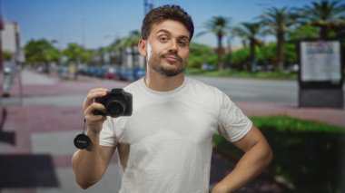 Güneşli bir şehir sokağında kamerayı tutan genç İspanyol bir adam, objektifin kapağını tutarken kolları bükülmüş; şehir hobisi.