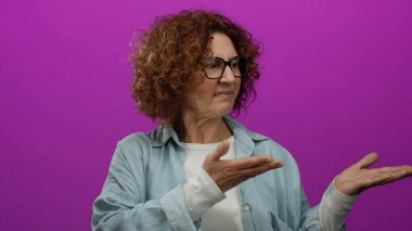 Elleri parlak pembe arka plana uzanmış, gözlük takan, kıvırcık saçlı ve tarafsız ifadeli mavi gömlekli bir kadın.