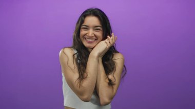 Gözleri kapalı gülümseyen İspanyol kadın, mor stüdyo arkaplanına karşı elleri yanaklarına kenetlenmiş; mutluluk sıcaklığı.