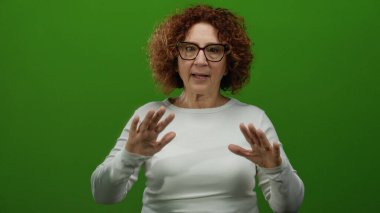 Katı yeşil duvar arka planına sakince el hareketleriyle iletişim ve sükunetini ifade eden ve rahat bir ortamda gözlük takan bir kadın..