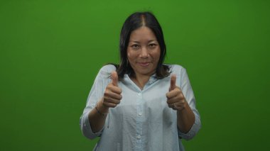 Açık mavi gömlek giyen, gülümseyen ve elini uzatan bir kadın yeşil stüdyoda kamerayı işaret ediyor..