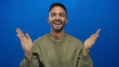 Elleri mavi bir arkaplan üzerinde gülümseyen genç bir adam, mutluluk ve açıklık kavramları için ideal, anlamlı bir jest olarak sevinç ve pozitifliği iletiyor..