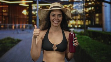 Hasır şapkalı kadın gülümsüyor ve kalabalık bir gece sokağında meyve suyu şişesini işaret ediyor; coşku.