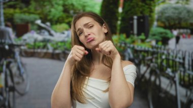 Surat asan dudakları ve yumrukları olan bir kadın, beyaz elbiseli, park halindeki bisikletli bir caddede yanaklara yakın duruyormuş. Üzüntü..