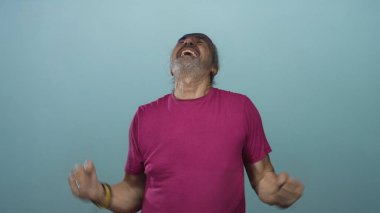 Beyaz uzun saçlı, sıkılmış yumrukları ve çıplak kolları olan bir adam, eflatun gömlekli, mavi bir duvarın önünde stüdyoda bağırıyor. Öfke boşalıyor..