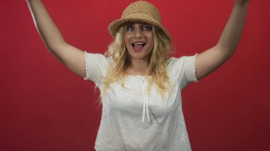 Hasır şapkalı ve beyaz bluzlu bir kadın kollarını başının üstüne kaldırıyor ve stüdyoda kırmızı arka planla gülümsüyor..