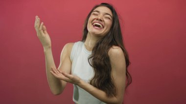 Gülümseyen genç İspanyol kadın pembe stüdyo ortamında rahat durarak el hareketi yapıyor; keyif daveti.