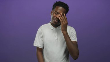 Beyaz polo tişört giyen koyu tenli bir adam mor stüdyoda diş ağrısı için yanağını tutuyor..