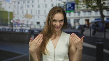 Beyaz kolsuz bluzlu kadın, yoğun şehir caddesinde gün ışığında elleri yanında seni çağırıyor; hoş geldin oyunculuğu daveti.