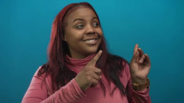 Pembe balıkçı yaka kazak giyen, kendine güvenen bir gülümsemesi olan bir kadın, bir deniz mavisi atölyesini işaret ediyor ve yan bakıyor..