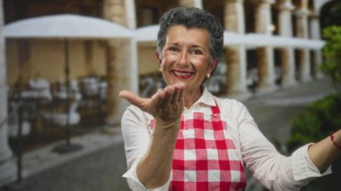 Gri saçlı ve önlüklü yaşlı kadın, açık kafeleri ve kemerleri olan eski bir kasaba sokağında, davetkar bir jest yaparak elini uzatıyor..