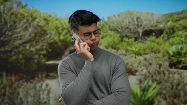 Parkta telefonla konuşan, gözlüklü yakışıklı bir adam. Çevresi yemyeşil, doğa ve modern iletişim ile huzurlu bir bağ sergiliyor..