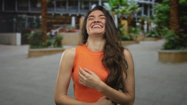 Turuncu tişörtlü kadın, palmiye ağaçlarının arasına cepheyi kurarak sokakta el ele gülüyor; kaygısız neşe.