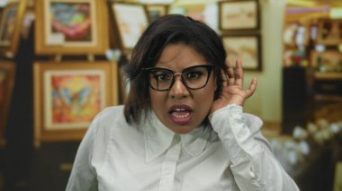 Sanat galerisinde dikkatle dinleyen, gözlüklü ve beyaz gömlekli, odaklanma ve merakı vurgulayan renkli resimlerle çevrili bir kadın..