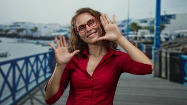 Kırmızı tişörtlü ve gözlüklü gülümseyen kadın sahildeki mavi korkulukların ve suyun arka planında neşeli bir tavırla geziniyor..