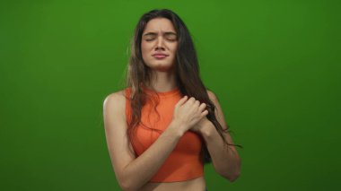 Genç İspanyol kadın ayakta duruyor ve göğsünü sıkıca tutuyor gözleri kapalı ve yeşil stüdyoda irkiliyor; acı.