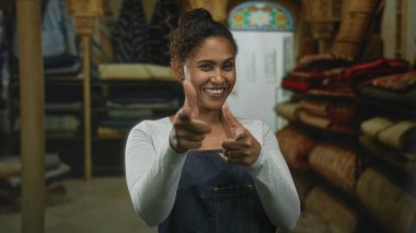 Kadın, bir inşaat atölyesinde yığılmış halı ve sürgüler arasında kamerayı işaret ediyor; dostça bir hizmet.