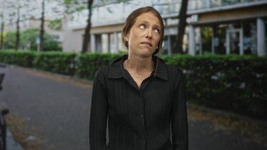 Kadın, bir binanın önündeki caddede dikilirken yüzünü ekşitiyor ve başını eğiyor, siyah bluz giyiyor, rahatsızlık veriyor..