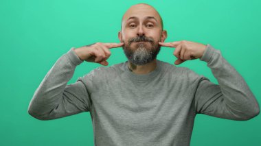 Sakallı kel adam izole edilmiş yeşil bir duvarın önünde neşeli tavırlarını ve günlük tarzını vurgulayarak neşeli ifadeler sergiliyor..