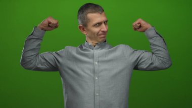 Yeşil arka plan önünde kaslarını esneten kendinden emin bir adam. İçeriden izole edilmiş gri gömlekli bir gülümsemeyle güç ve kararlılık gösteriyor.