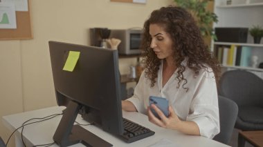 Ofis binasındaki bilgisayar monitörüne bakarken elinde akıllı telefon tutan kadın, verimliliğe odaklanma.