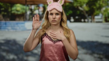 Pembe gece elbisesi ve uyku maskesi takmış genç bir kadın, elleri göğsünde, gün ışığında durgun bir poz veren yeşil ağaçlarla parkta duruyor..