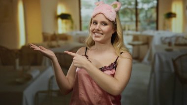 Pembe gecelikli bir kadın restoranda masalarla gülümsüyor, neşeli bir ifadeyle işaret ediyor ve sevimli bir uyku maskesi takıyor..