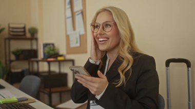 Gözlüklü genç sarışın kadın ofisteki akıllı telefonunu zevkle görüyor. Heyecan ve mutluluk taşıyor..
