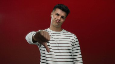 Çizgili gömlekli genç adam, izole edilmiş kırmızı arka planda baş parmaklarını aşağı gösteren jestler gösteriyor. Onaylamamanın ve memnuniyetsizliğin güçlü bir ifadesi..